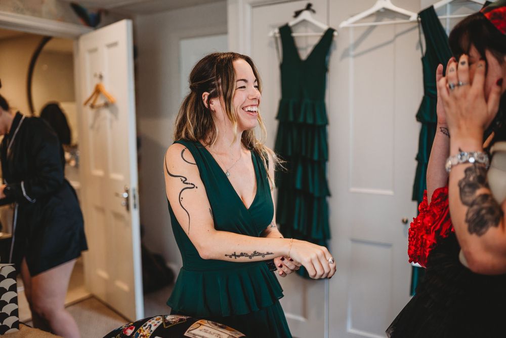 Woman in emerald green bridesmaid dress laughing during wedding preparations in a dressing room.