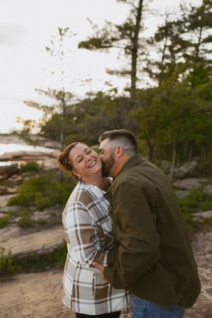 Man in green jacket kissing woman in plaid shirt on the cheek outdoors, both laughing near a rocky forest shoreline.