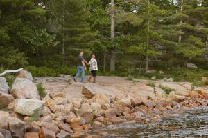 Couple walking hand in hand along a rocky lakeside trail surrounded by lush green pine trees in nature.