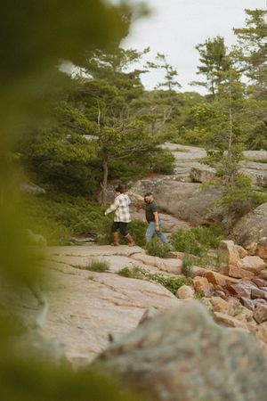Two hikers walking on rocky terrain surrounded by lush green trees in a scenic outdoor landscape.