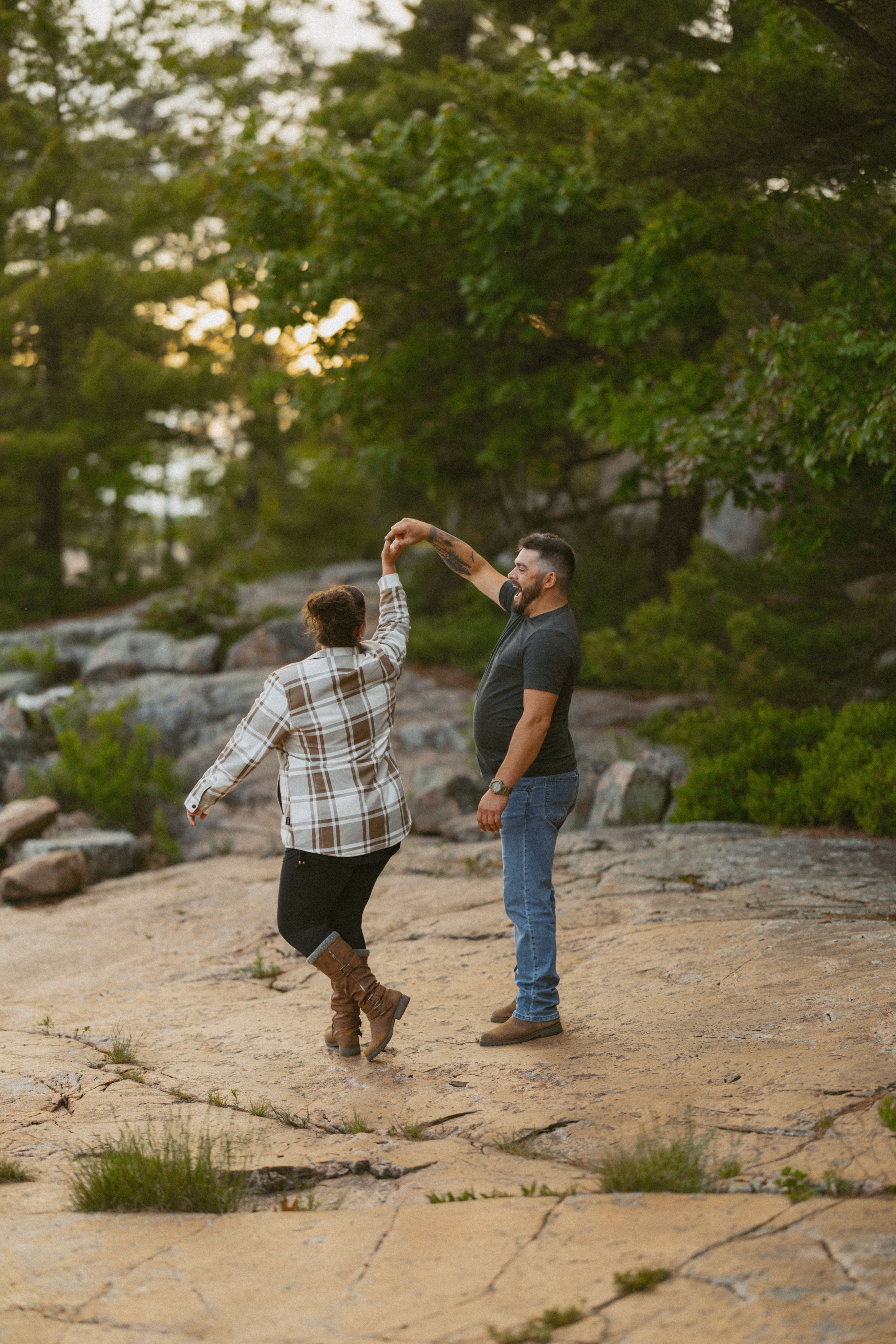 Couple dancing together on rocky terrain surrounded by lush green trees at sunset.