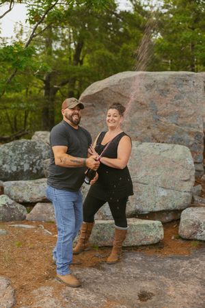 Couple laughing while popping champagne bottle outdoors near large rocks and green trees.
