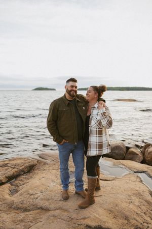 Couple standing on rocky shoreline by a lake, wearing casual fall outfits, smiling at each other with islands in background.