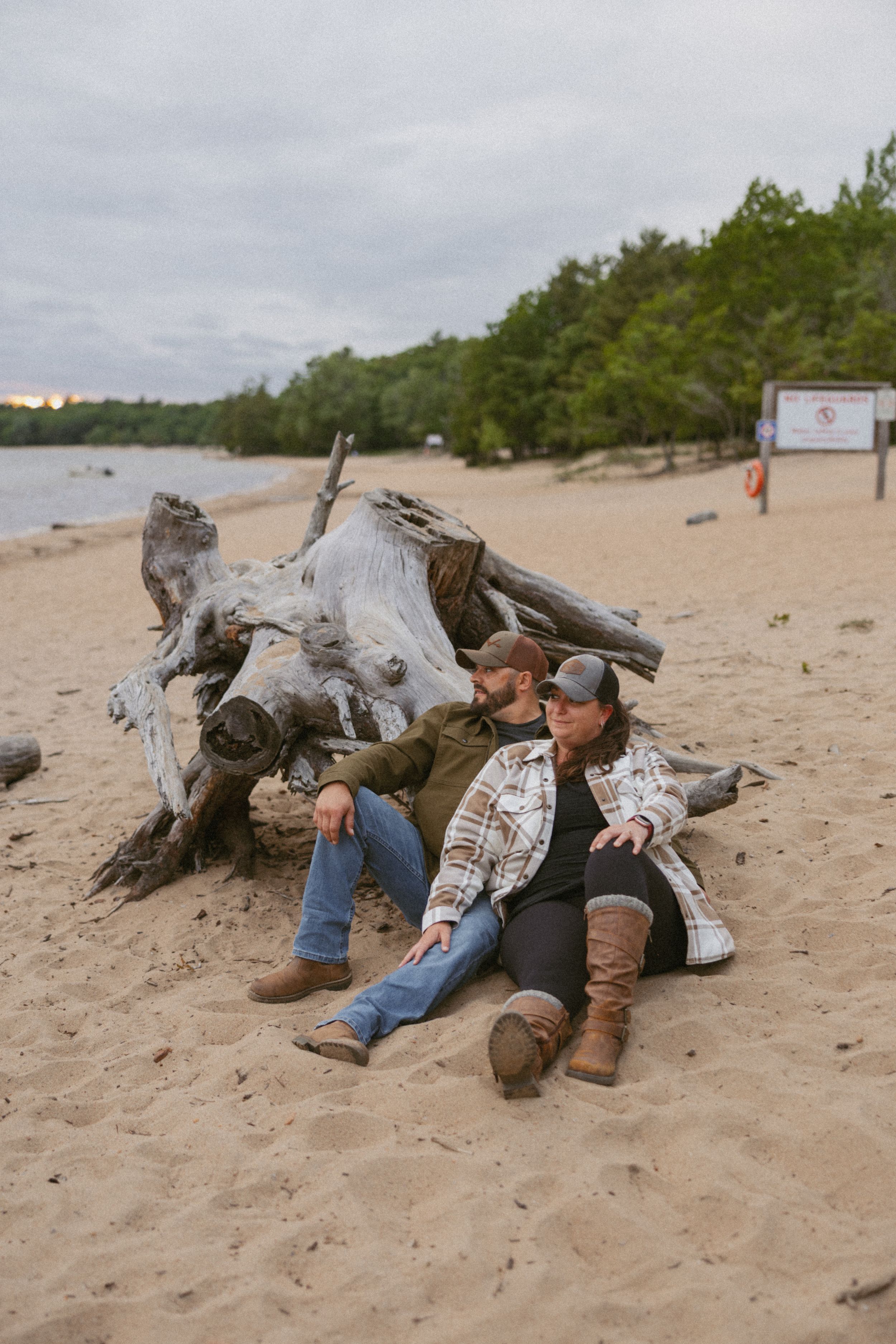 Couple relaxing against driftwood on a sandy beach, wearing flannel shirts and caps on a cloudy day.