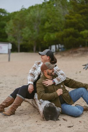 Couple sitting on driftwood log on sandy beach, wearing flannel shirts and caps, surrounded by green trees.