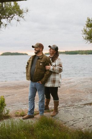 Couple in Carhartt jackets and caps standing on rocky lakeside at sunset, embracing outdoors.
