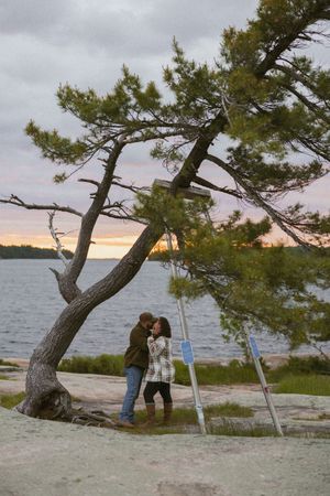 Couple sharing a kiss under a leaning pine tree by a lake at sunset on rocky shore.