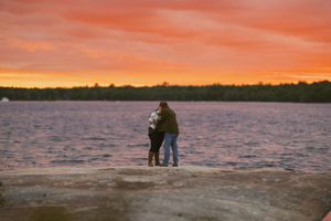 Couple embracing on rocky shore at sunset with vibrant orange sky reflecting over calm lake and tree-lined horizon.