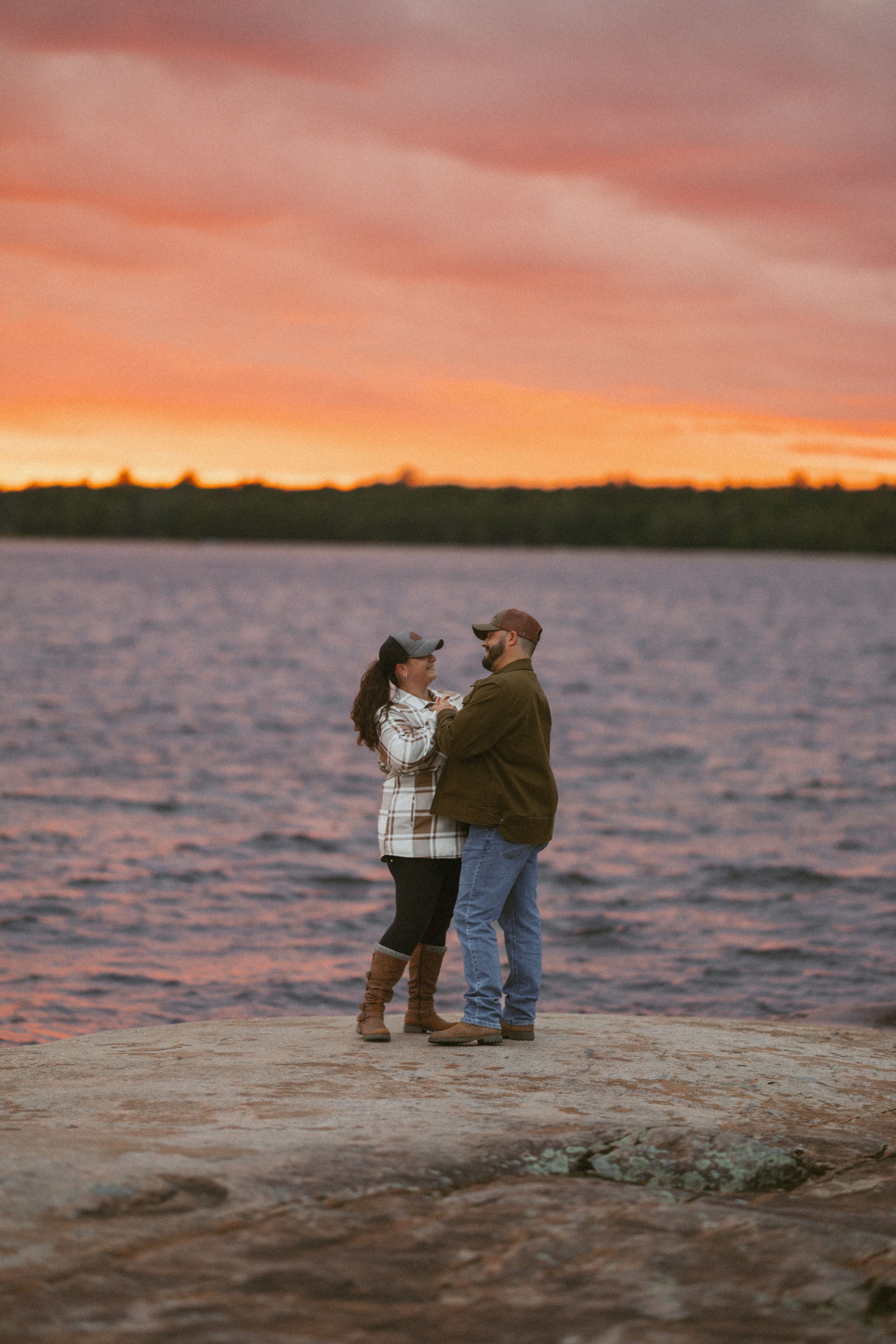 Couple embracing on rocky lakeside under a vibrant pink and orange sunset sky.