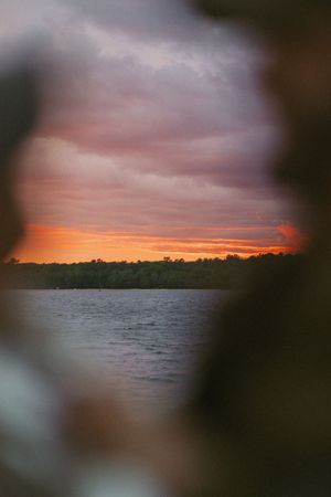 A stunning sunset viewed through foliage, with vivid orange sky over a calm lake and tree-lined horizon.