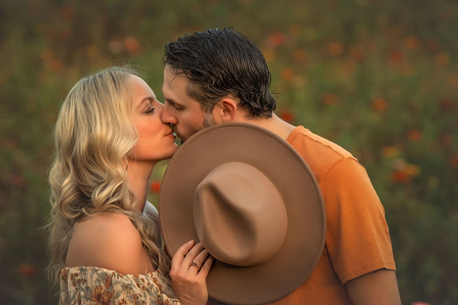 A couple sharing a romantic kiss in a flower field, the woman holding a tan wide-brim hat.