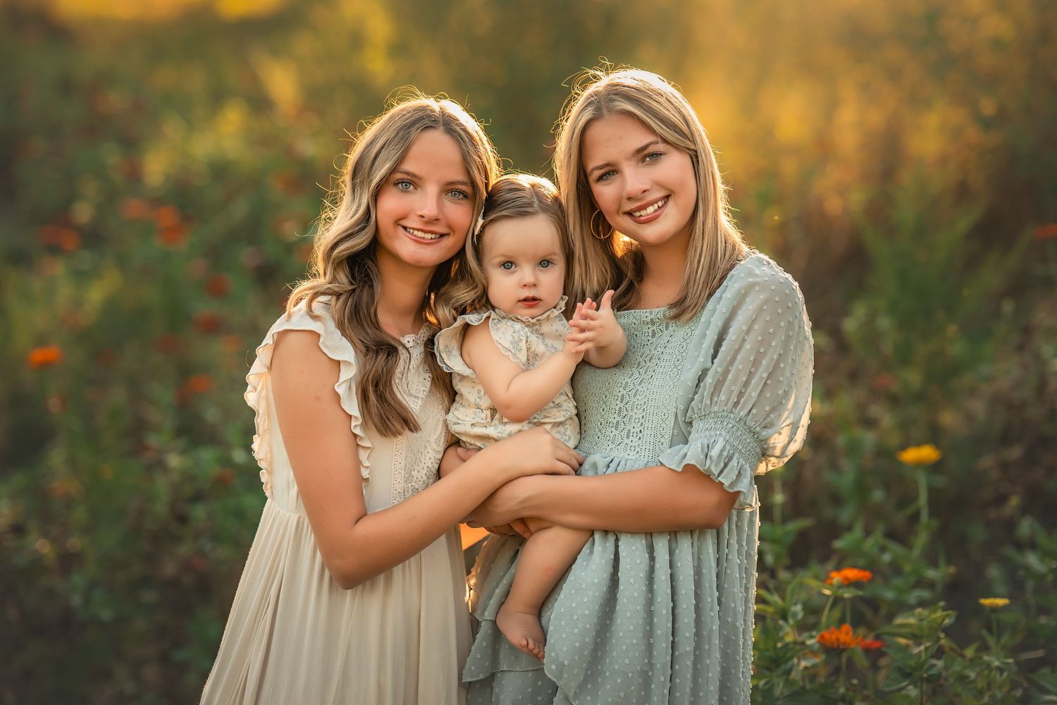 Two blonde twin sisters and a baby sister pose together in a sunlit flower field wearing soft pastel dresses.
