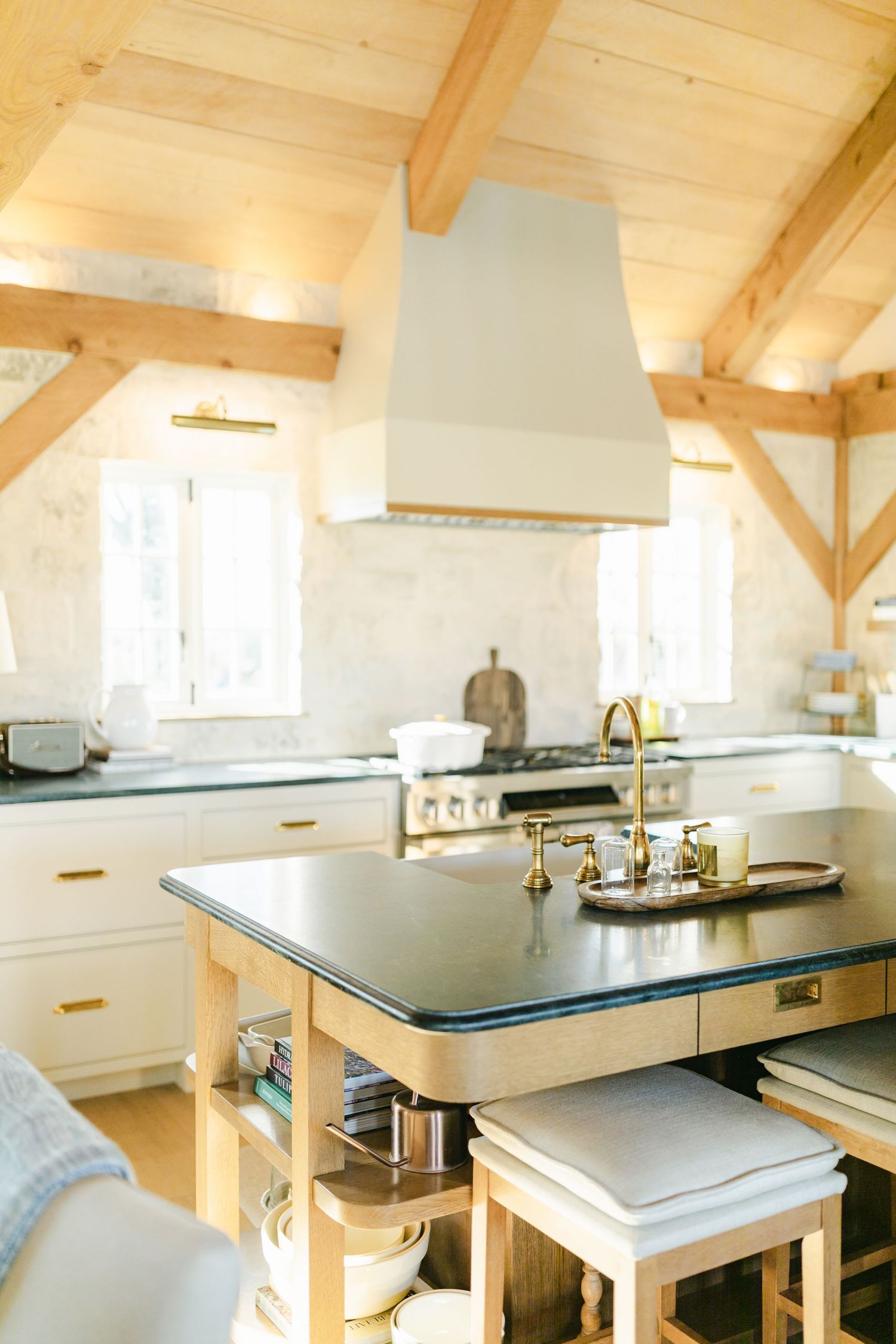 Bright farmhouse kitchen with wood beam ceiling, white cabinets, brass fixtures, dark granite island, and upholstered stools.