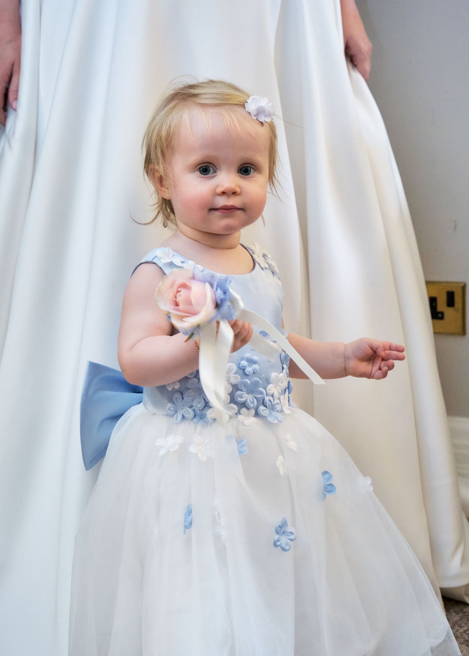 Adorable toddler girl in white and blue floral dress with bow and pink rose at a wedding.