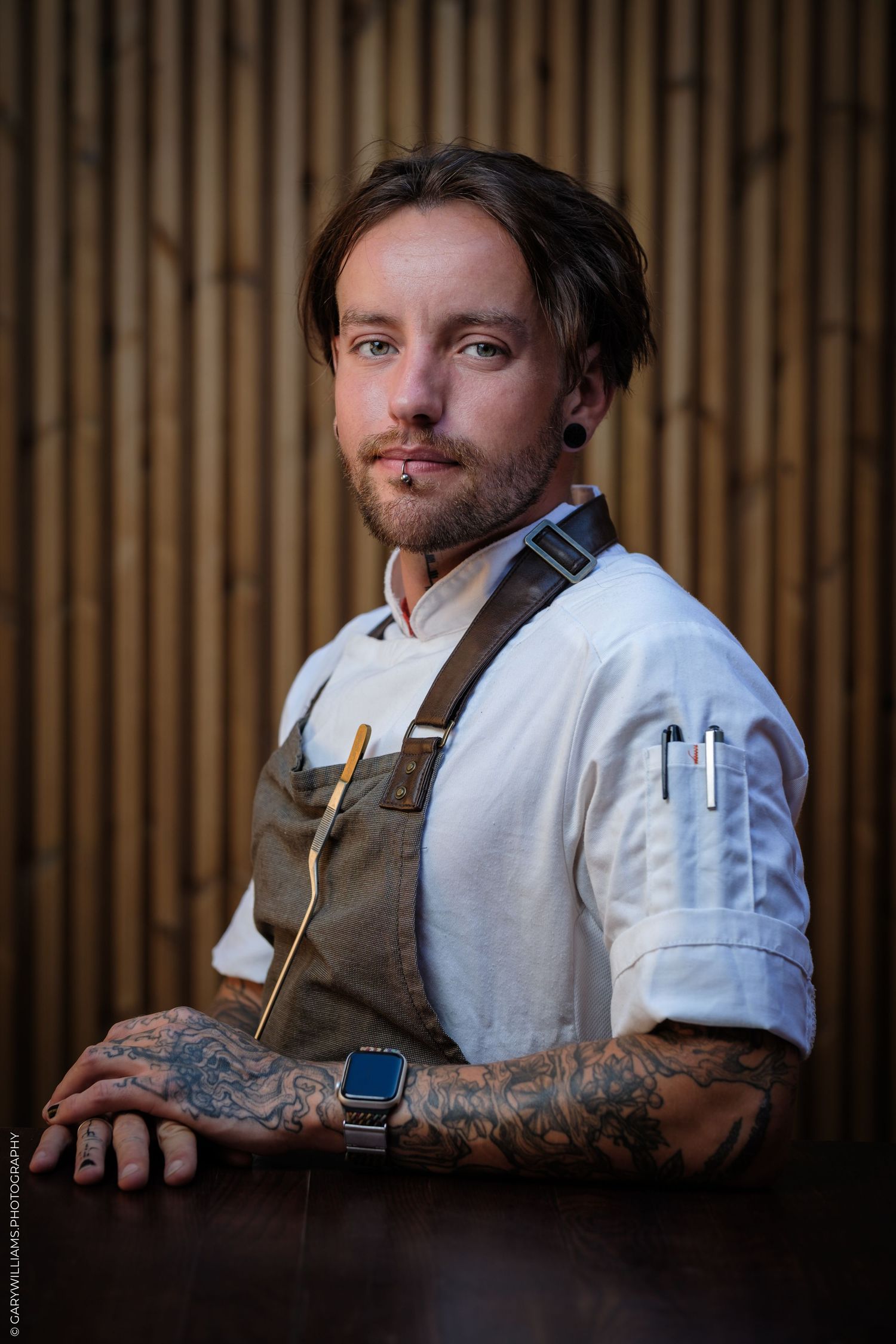 Tattooed chef in apron and white coat poses confidently with a spoon, showcasing culinary expertise.