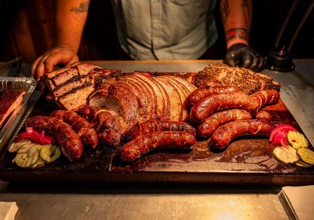 Large wooden board piled with smoked sausages, brisket, and BBQ meats with pickles and onions.