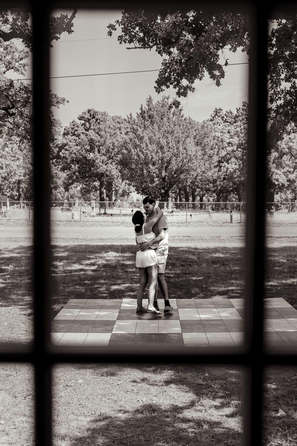 Black and white photo of couple embracing on checkered outdoor patio framed by window bars.