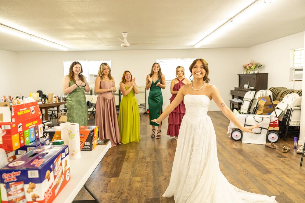 Bride in white gown smiling at bridesmaids in colorful dresses during wedding preparations indoors.