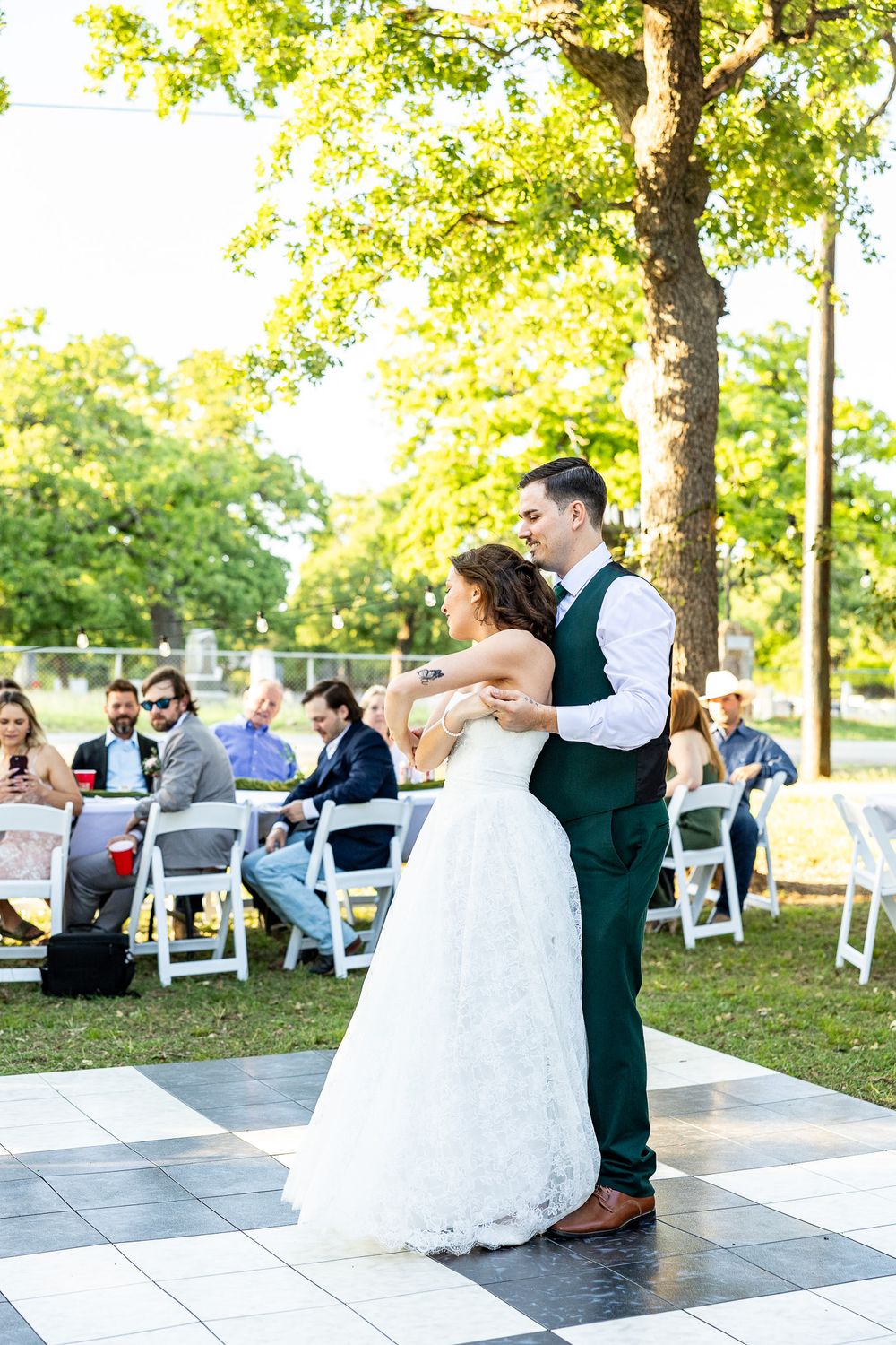 Bride and groom sharing first dance on checkered outdoor floor surrounded by seated wedding guests.