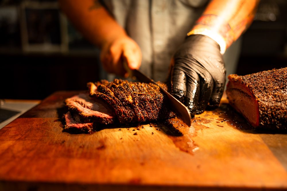 Person in black gloves slicing freshly smoked brisket on a wooden cutting board.