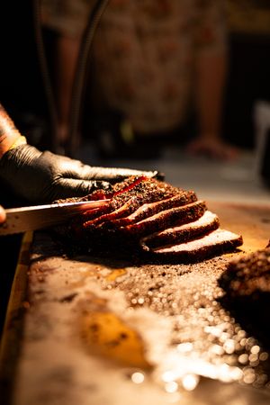 Pitmaster in black gloves slicing seasoned smoked brisket on a cutting board with a sharp knife.
