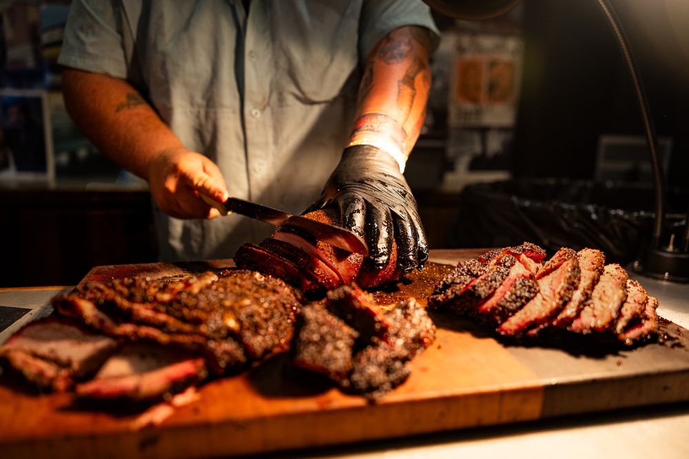 Chef in black gloves carving thick slices of smoked BBQ brisket on a wooden cutting board.