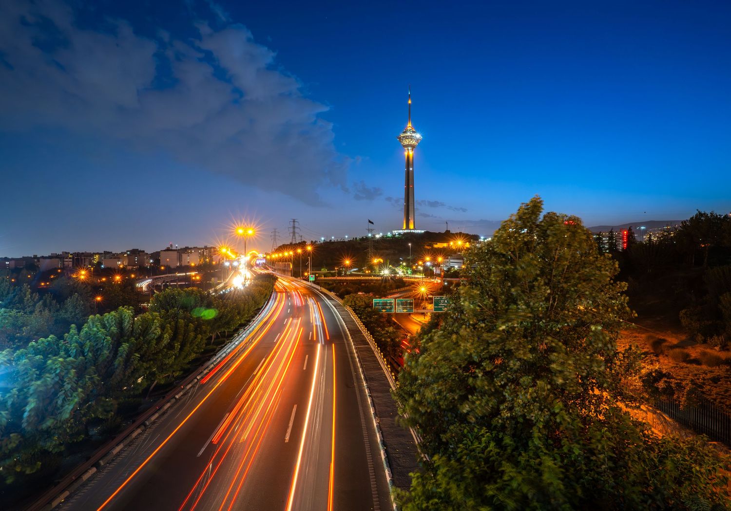 Milad Tower lit up at night in Tehran, with streaking light trails from highway traffic below.