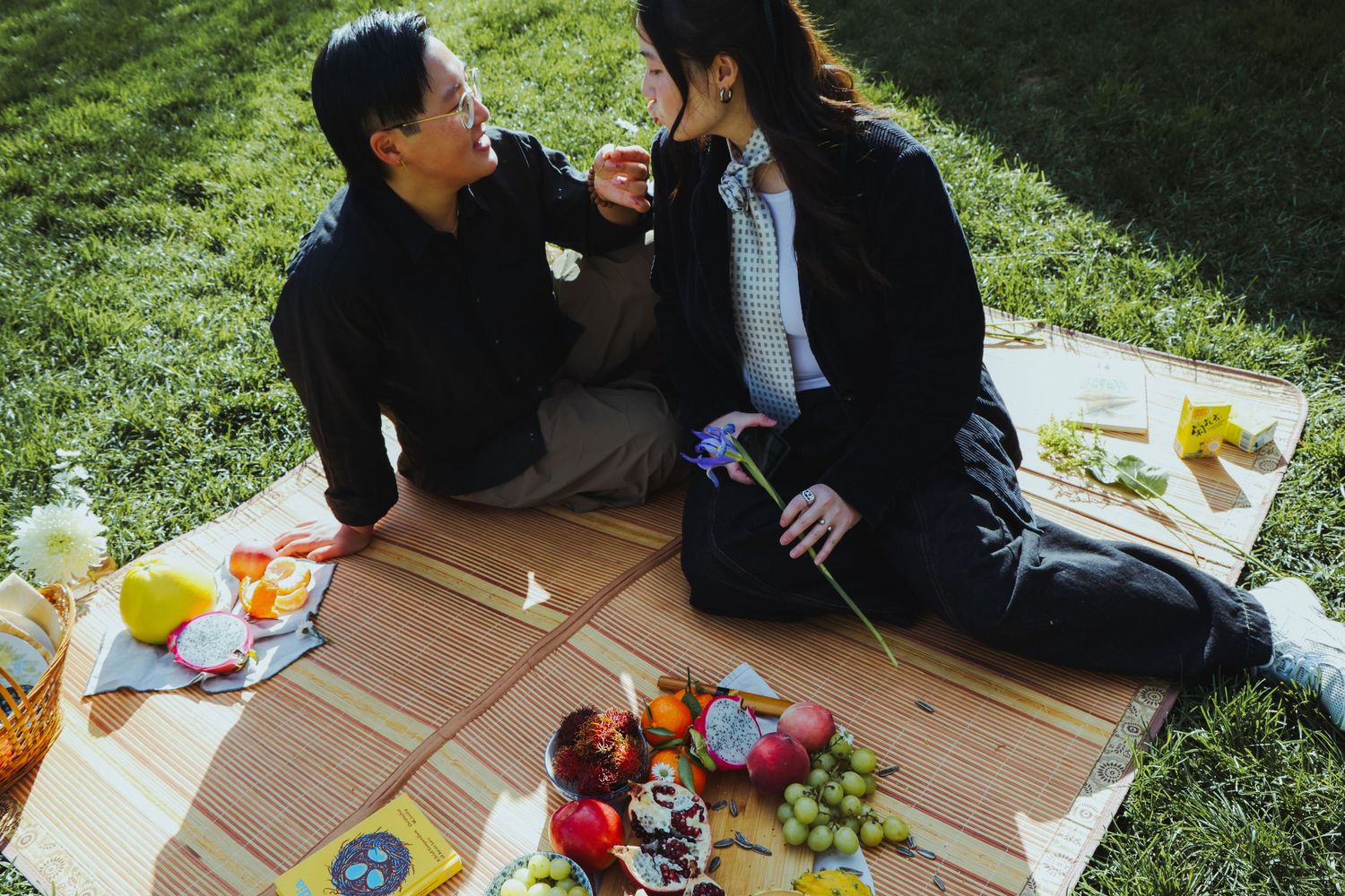 A couple picnicking in the grass, surrounded by different fruits and flowers at the Lewis MacAdams Riverfront Park in Los Angeles.