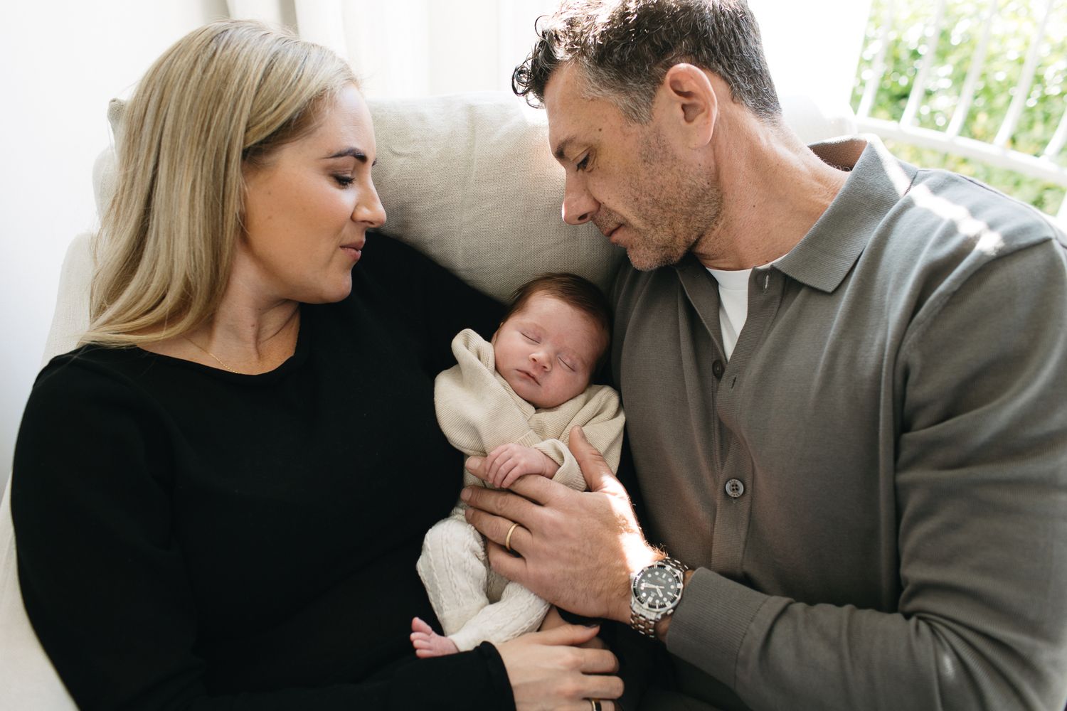 A couple tenderly gazes at their sleeping newborn baby wrapped in a white blanket in a bright, cozy room.