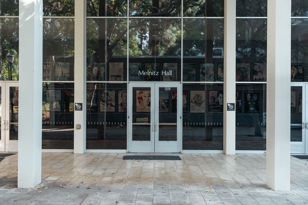 Exterior entrance of Meintz Hall featuring large glass doors and windows surrounded by white columns and lush trees.