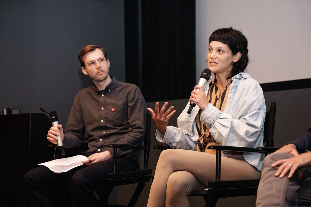 Two panelists speak at an indoor event, a man holding papers and a woman gesturing with a microphone.