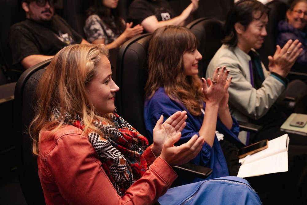 Audience members clapping and smiling at an indoor event, woman in red jacket and blue dress in foreground.