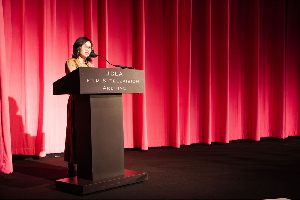Woman speaking at UCLA Film & Television Archive podium on stage with red curtain backdrop.