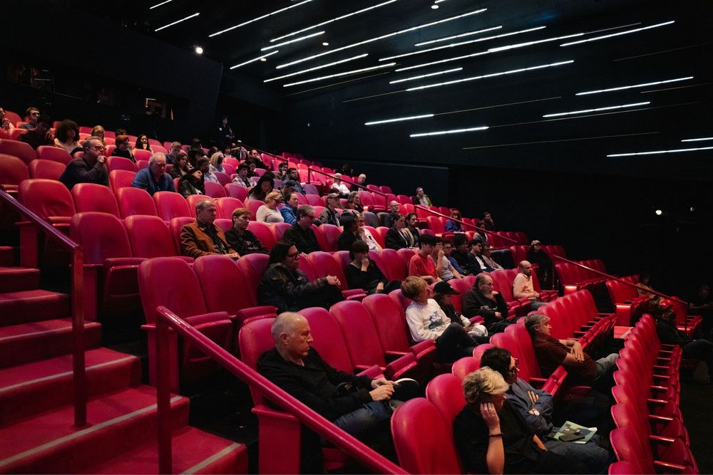Audience seated in red velvet chairs inside a modern cinema theater with dark walls and ambient lighting.