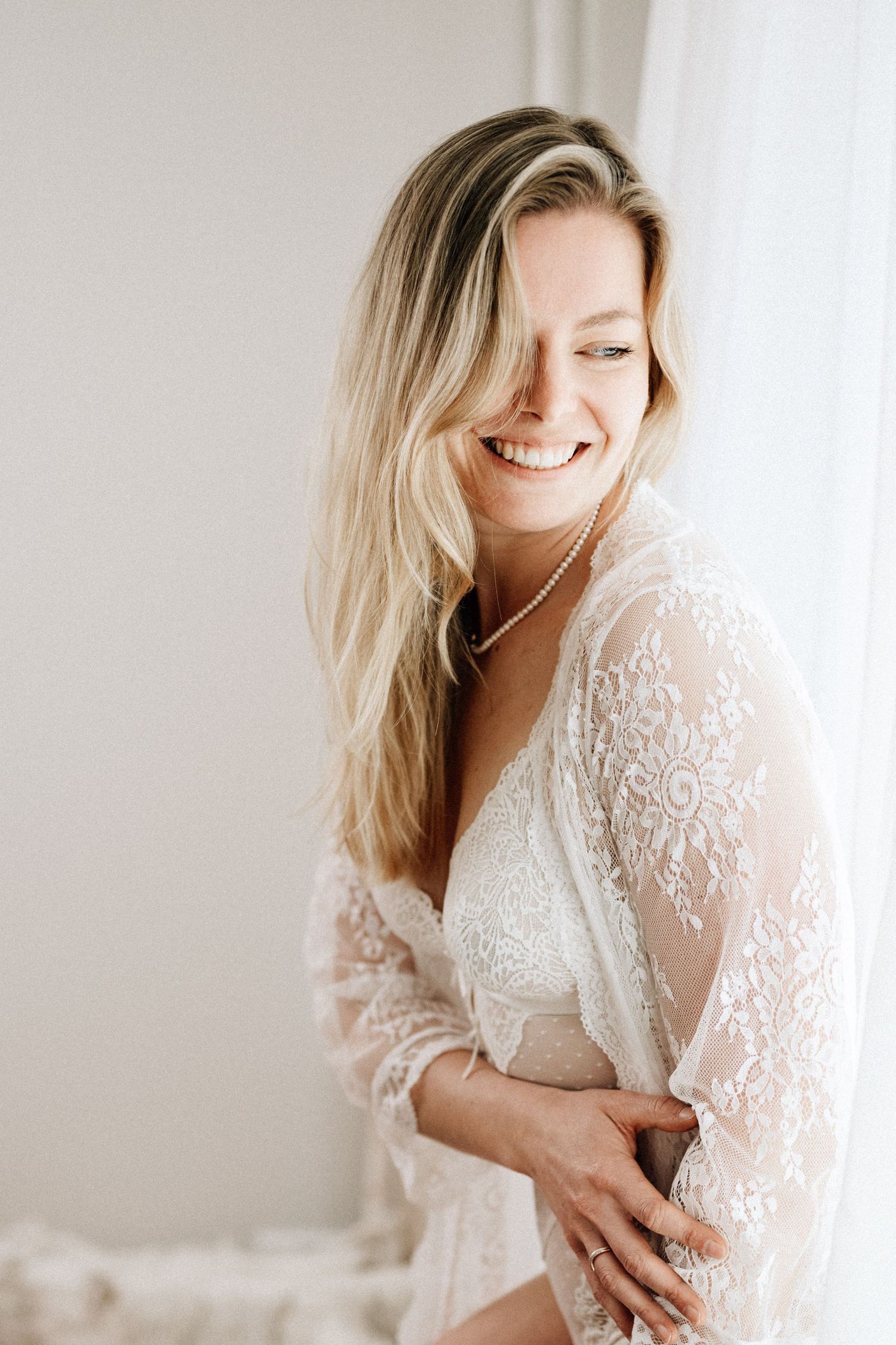 Boudoir Portrait einer Frau in einem Spitzencardigan stehend am Fenster mit sanftem Licht.