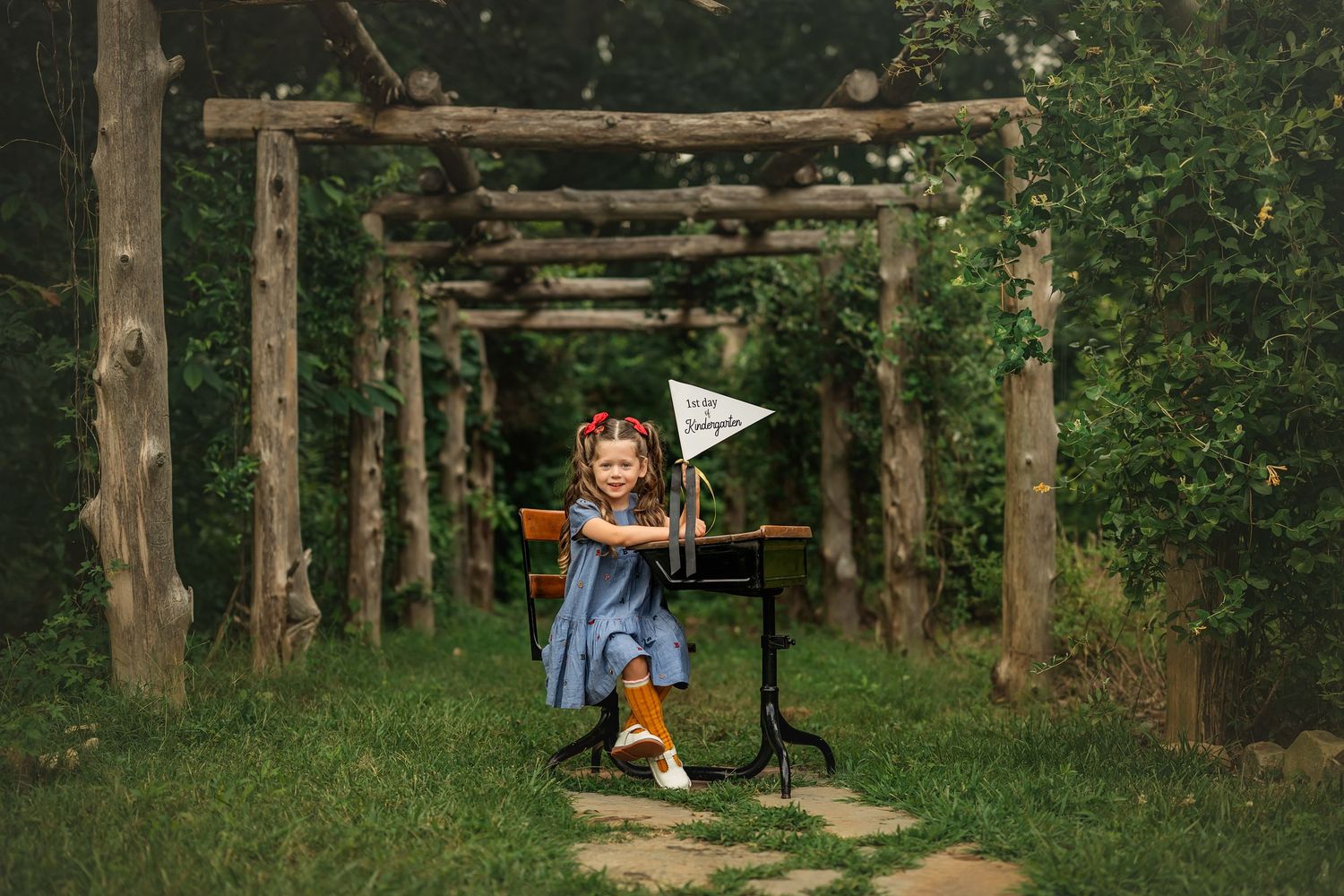 A young girl sits at a vintage desk outdoors under a wooden arbor, holding a pennant flag in a lush green garden.