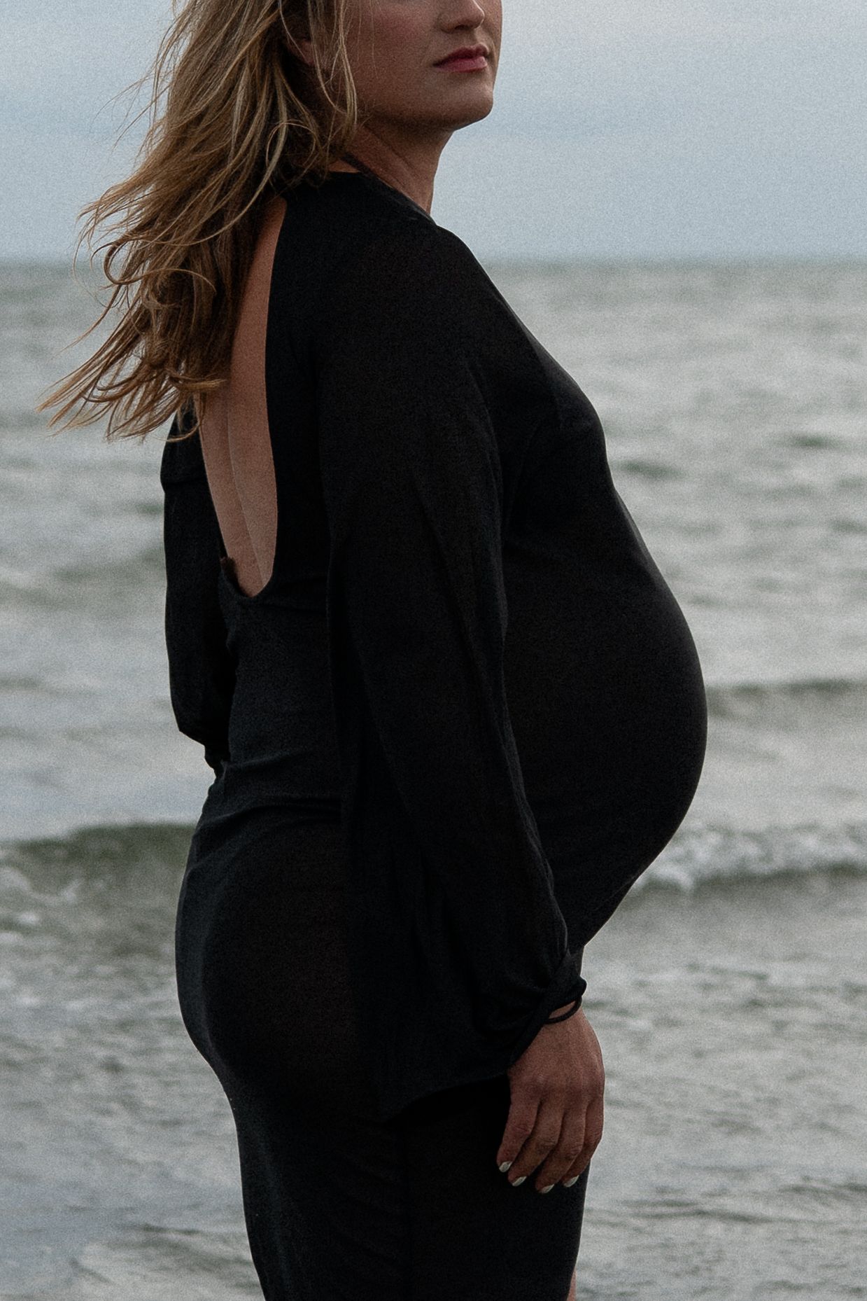 Pregnant woman in fitted black open-back dress posing sideways on overcast beach with ocean behind her.