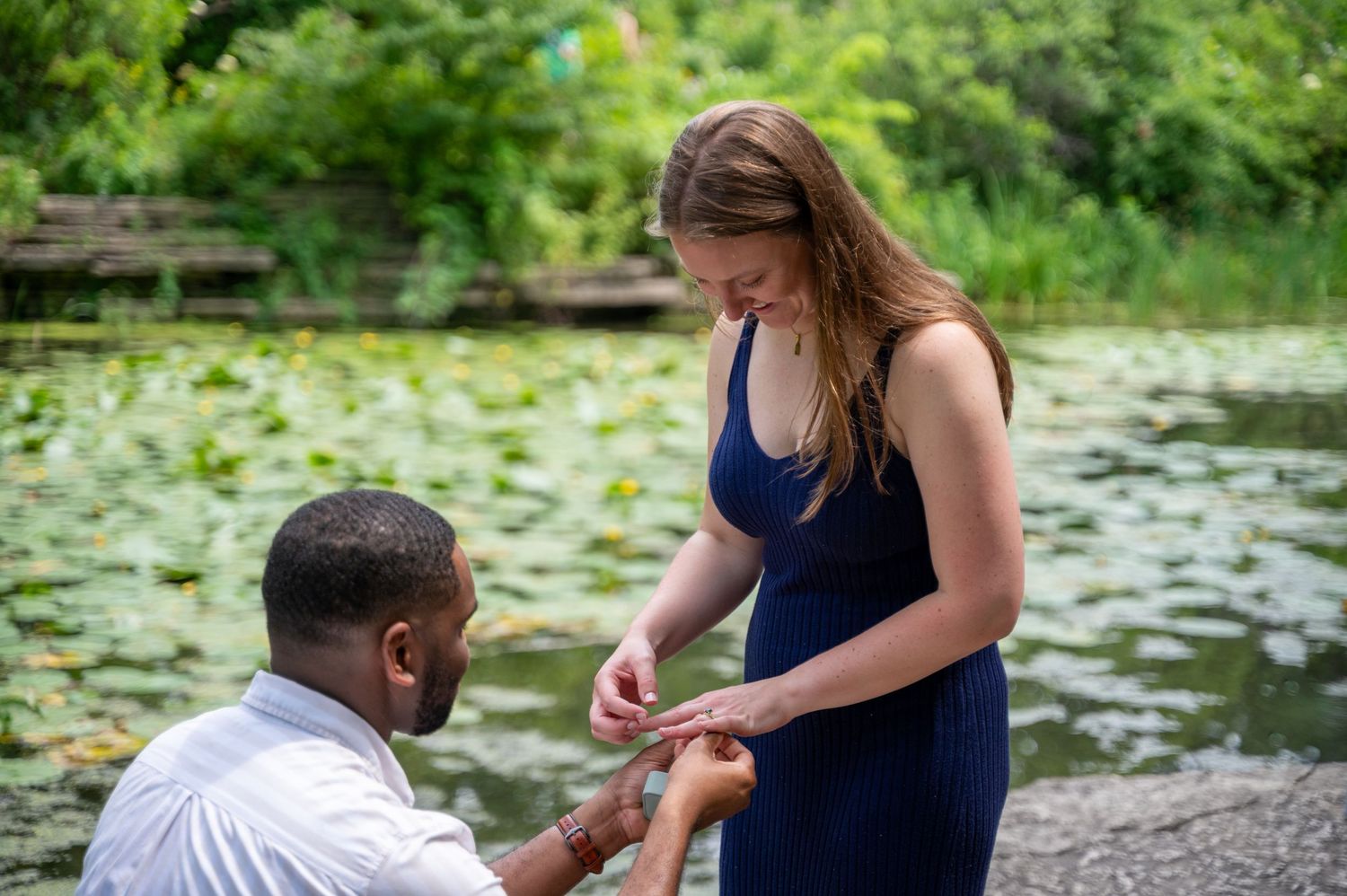 Man kneeling proposing to woman in blue dress by a serene lily pad pond outdoors.