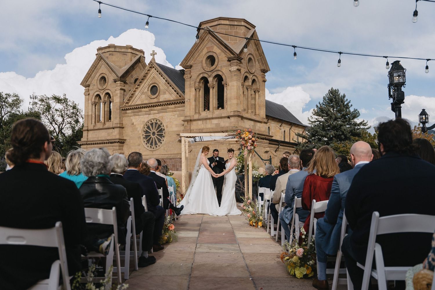 Outdoor wedding ceremony in front of a historic stone church in Santa Fe, with guests seated in white chairs.