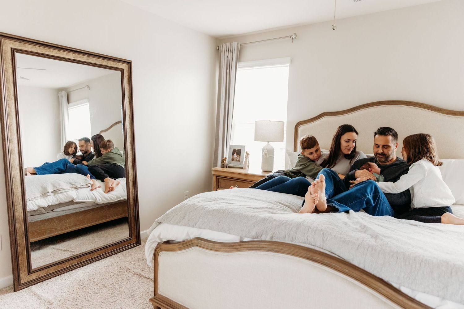 Family relaxing together on a bed in a bright bedroom with a large floor mirror reflecting their cozy moment.