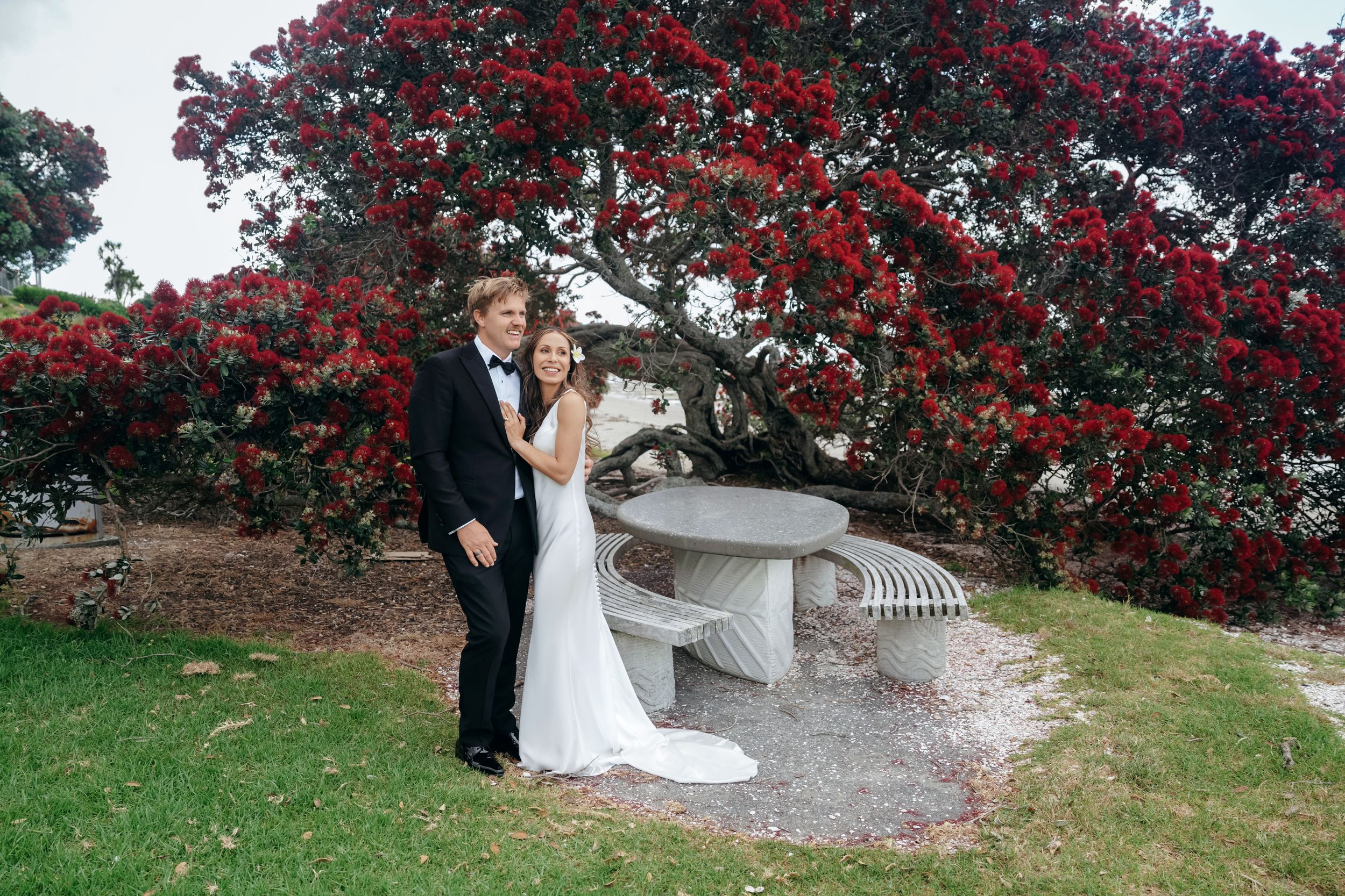 Bride and groom pose together under a vibrant red pohutukawa tree beside a stone bench on their wedding day.
