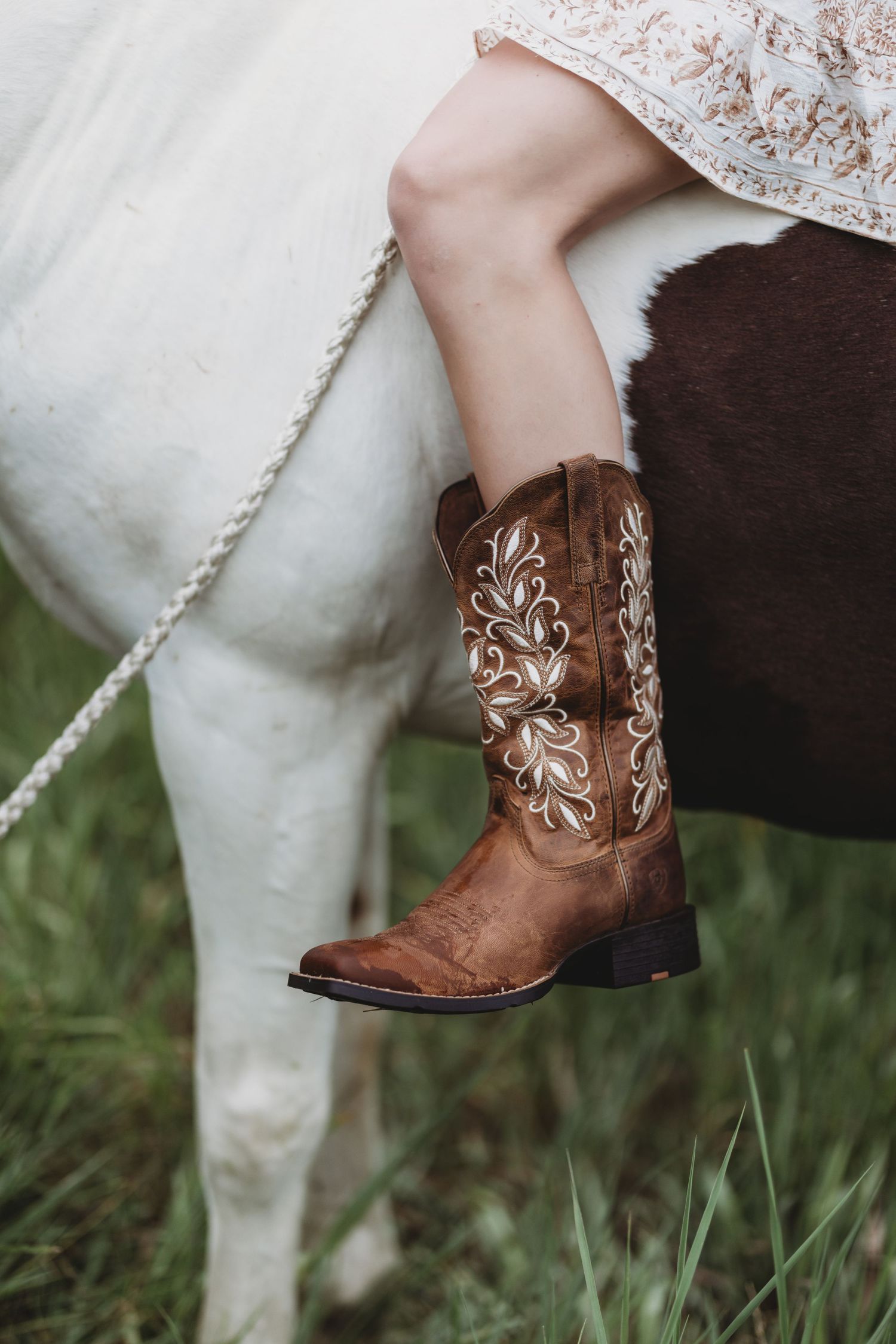 Woman wearing embroidered brown cowboy boots sitting on a horse in a grassy field.