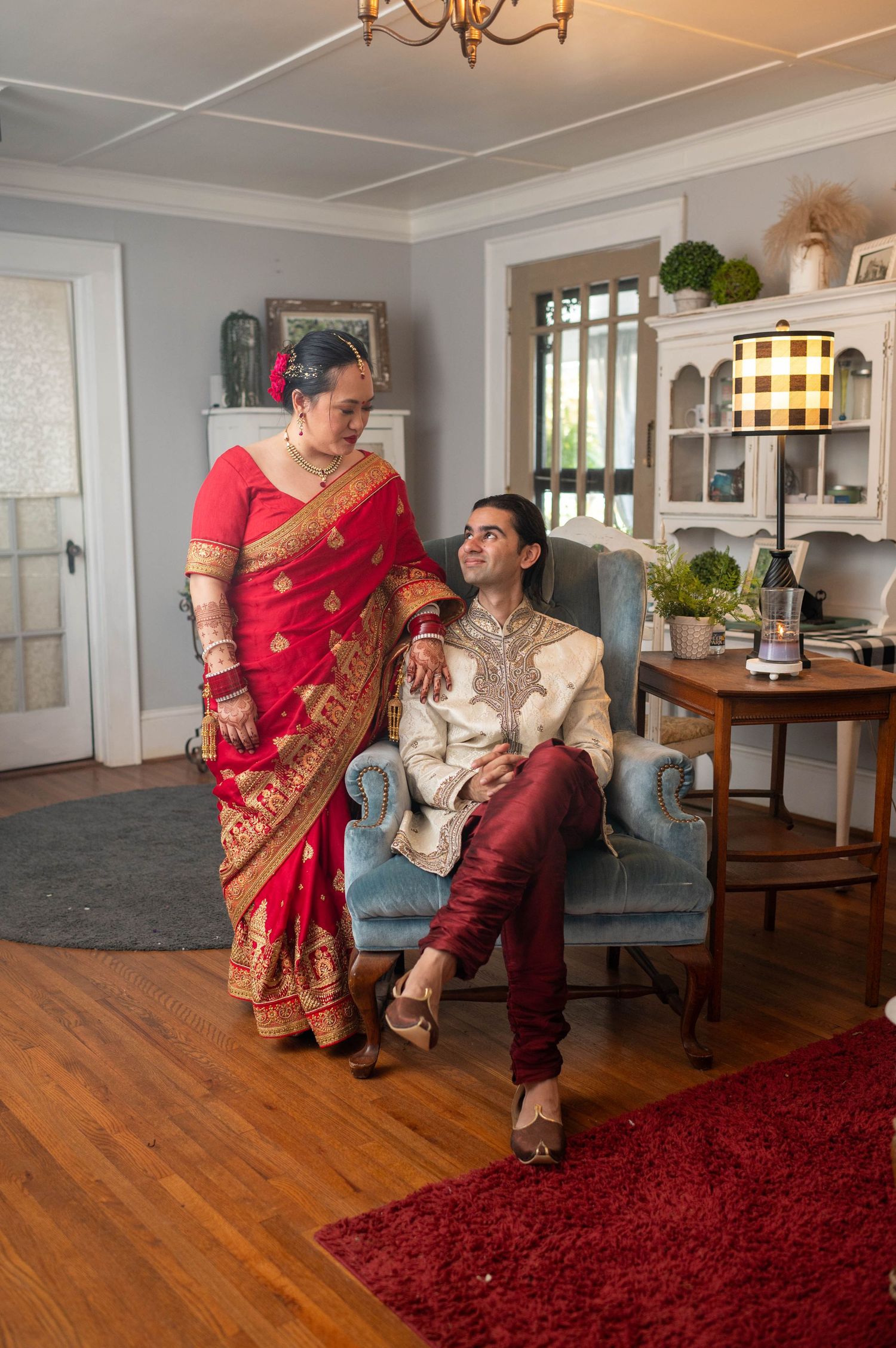 Couple in traditional Indian wedding attire, bride in red saree and groom in cream sherwani, posing in elegant room.