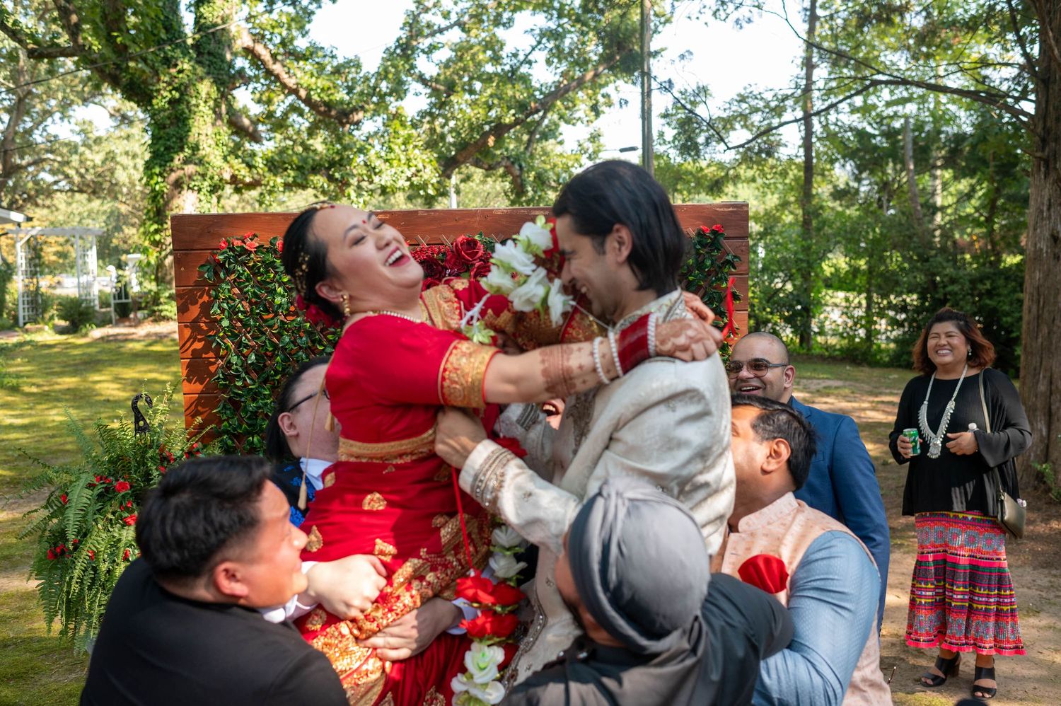Joyful South Asian wedding ceremony with bride in red lehenga and groom in gray sherwani embracing outdoors.
