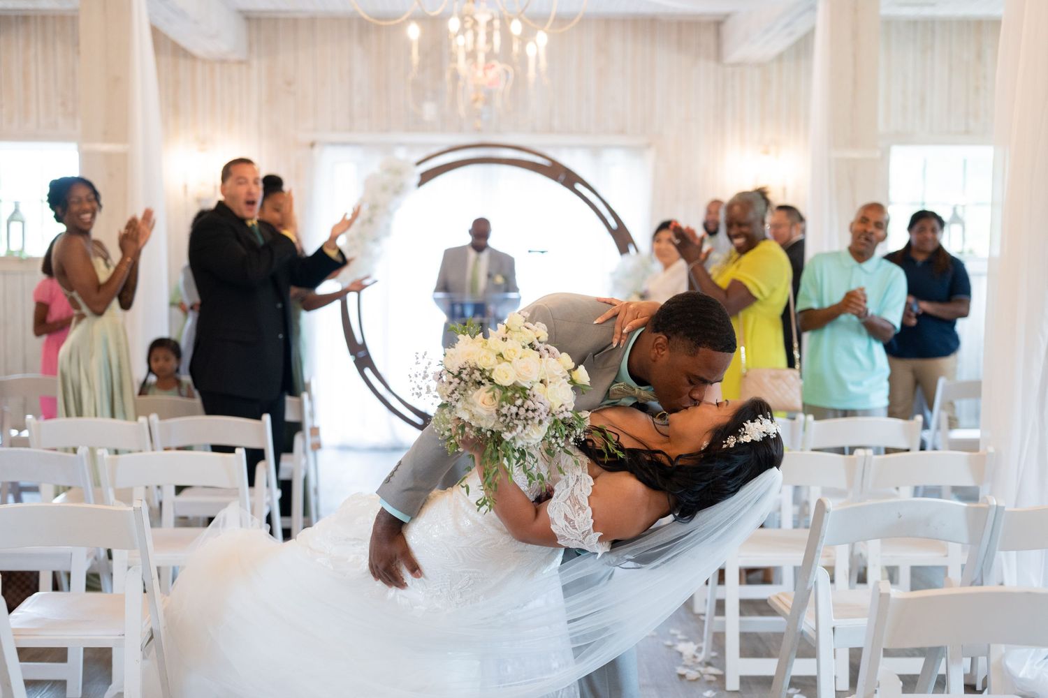 Groom dips bride for a kiss at wedding ceremony in elegant white chapel with guests applauding.