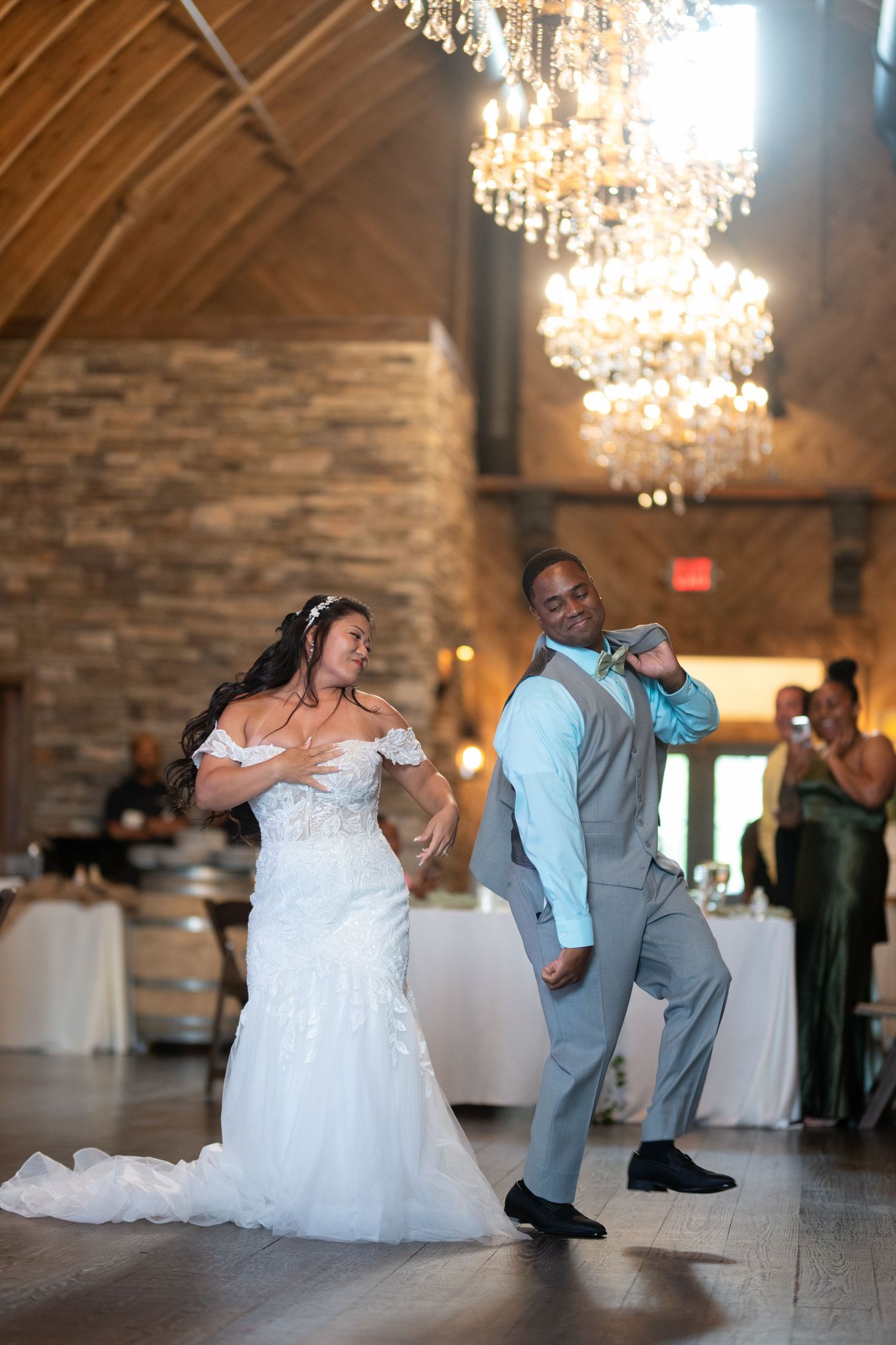 Bride and groom share a fun, energetic first dance in an elegant rustic barn wedding venue with crystal chandeliers.