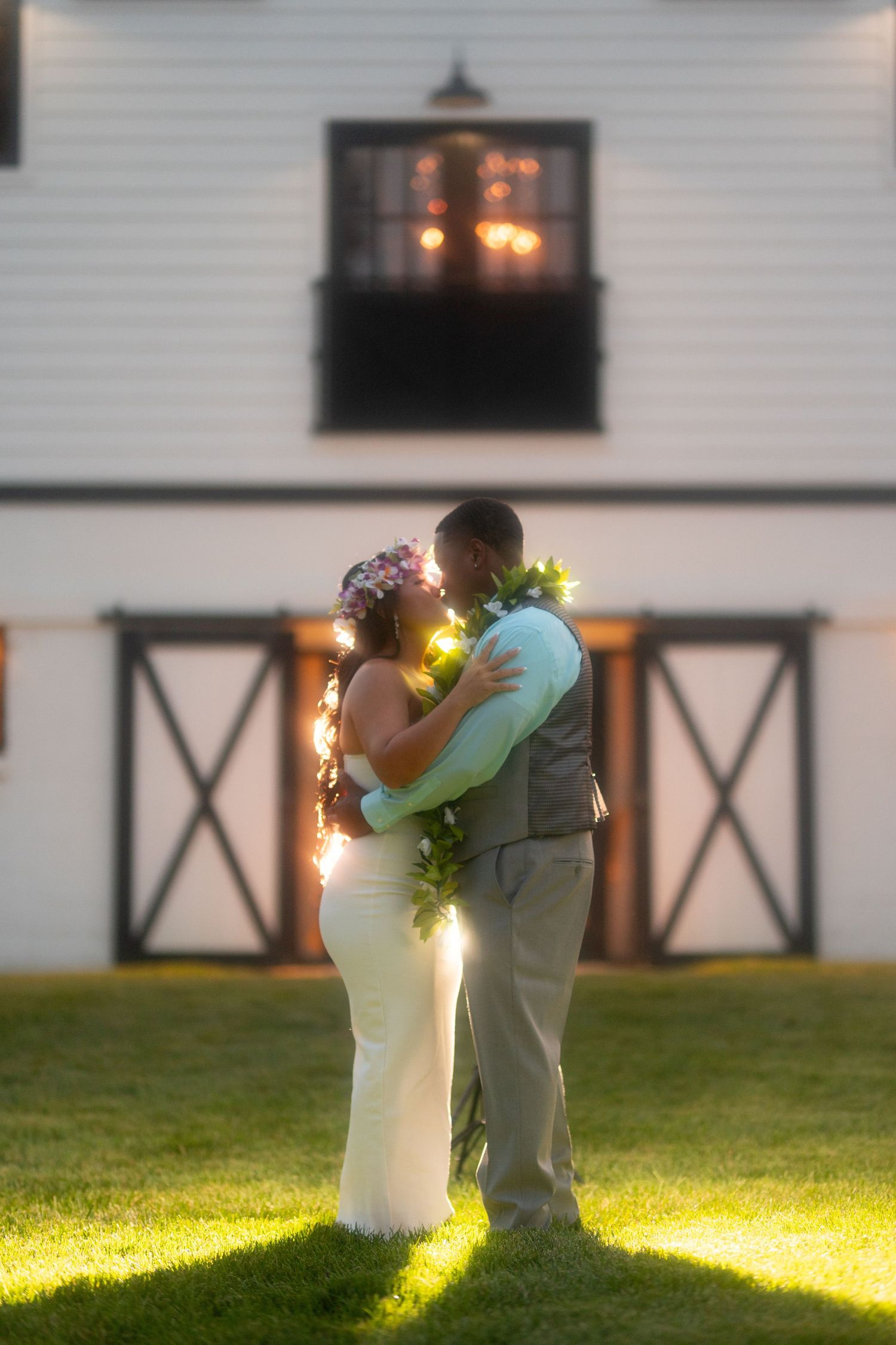 Bride and groom sharing a romantic kiss on a lawn at dusk, adorned with floral leis and fairy lights near a white barn.