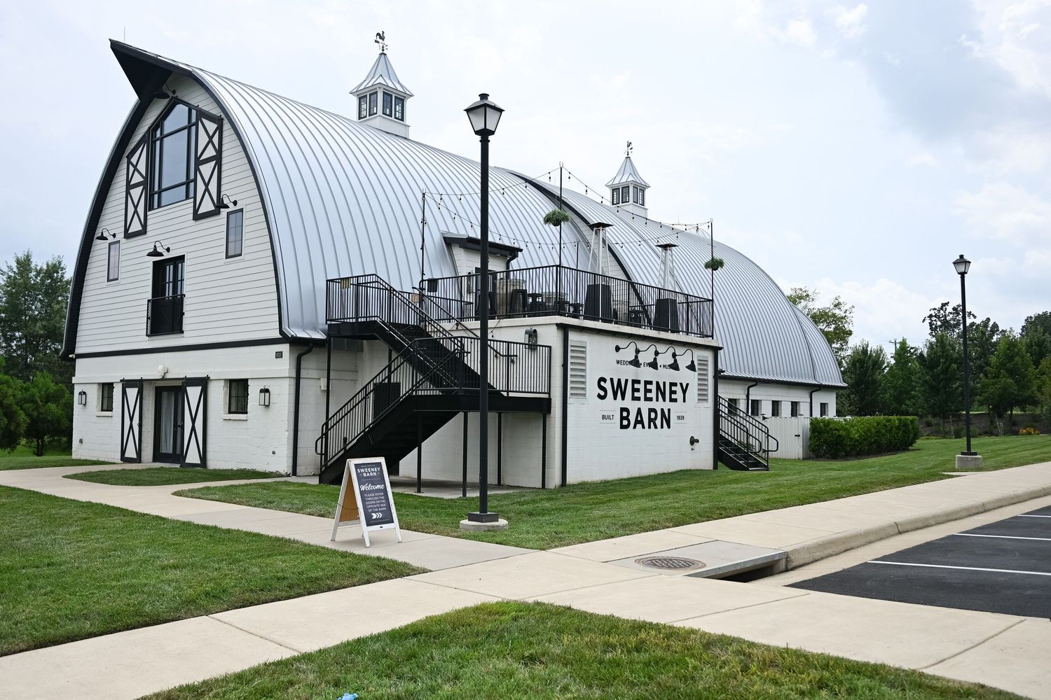 Sweeney Barn event venue exterior featuring a white gambrel-roof barn with black metal stairs and lamp posts on a sunny day.