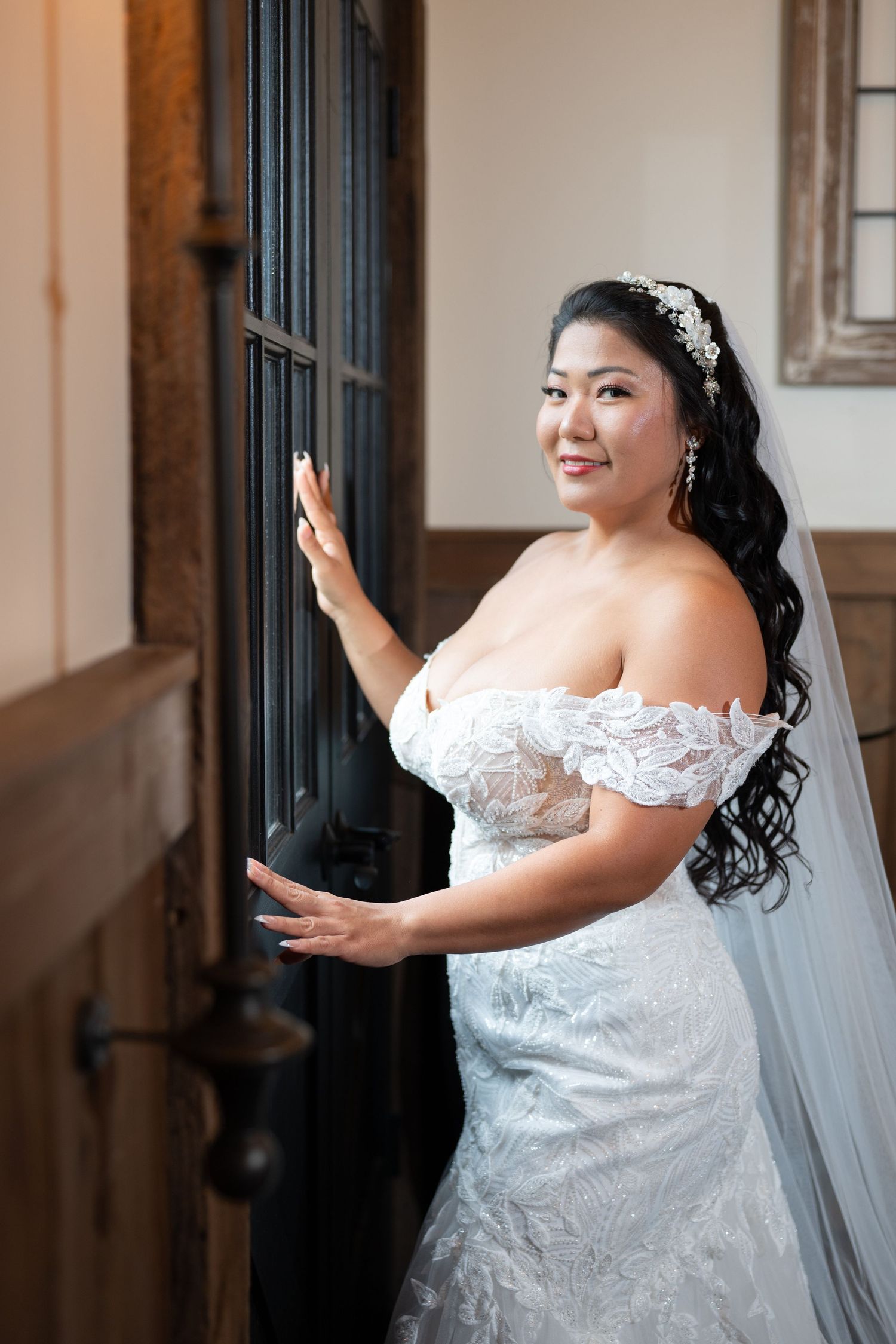 Bride in off-shoulder lace wedding gown and veil poses elegantly by ornate dark wooden door, smiling over shoulder.