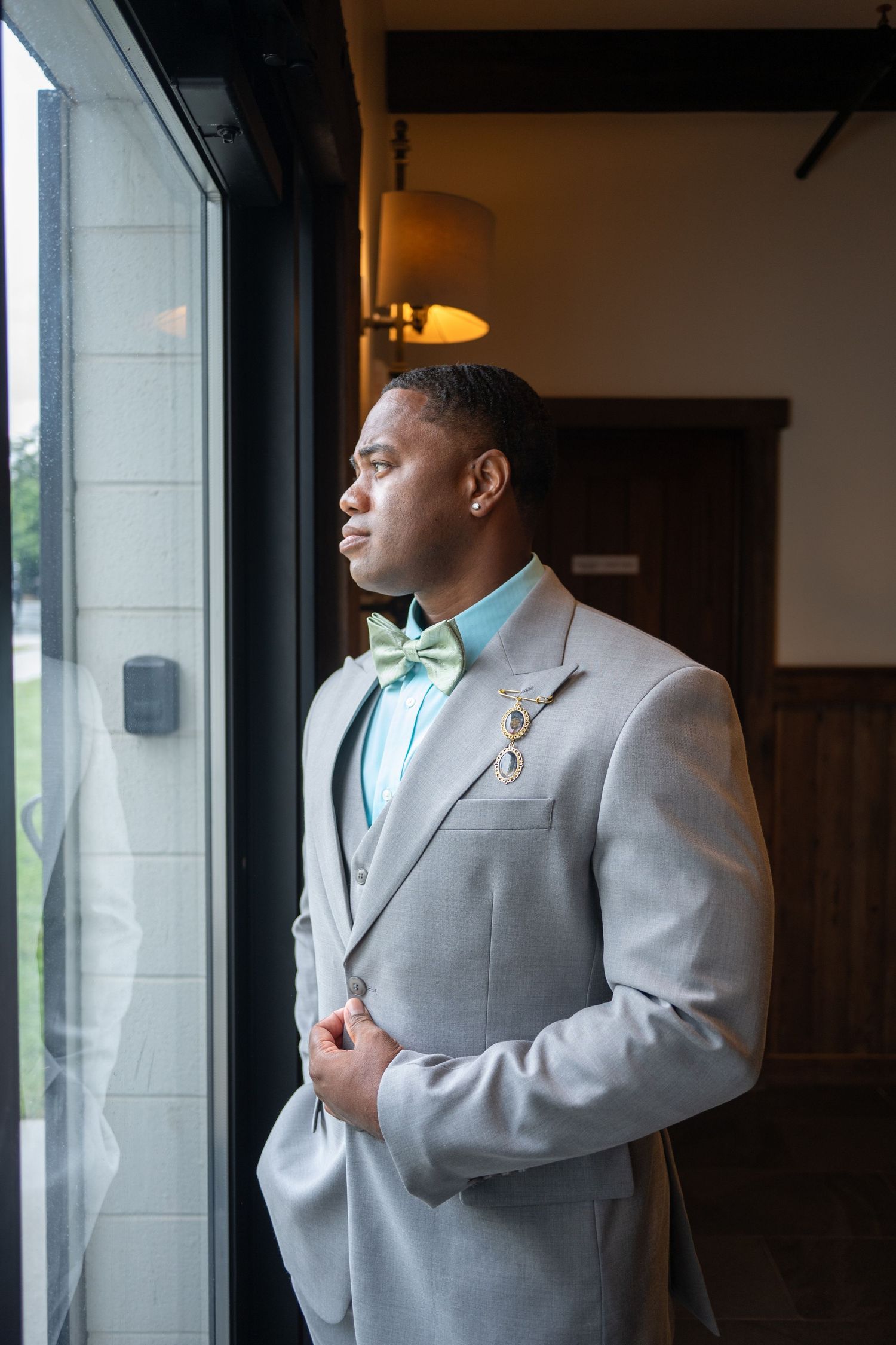 A well-dressed Black man in a gray suit with mint bow tie and boutonniere gazes out a window at a formal event.